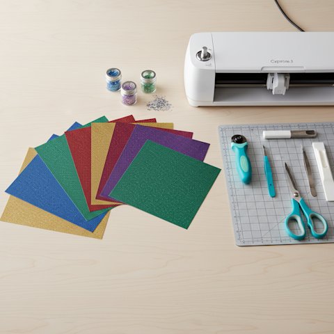 Flat-lay of jewel-tone glitter cardstock sheets fanned beside a Cricut machine and crafting tools on a wood desk